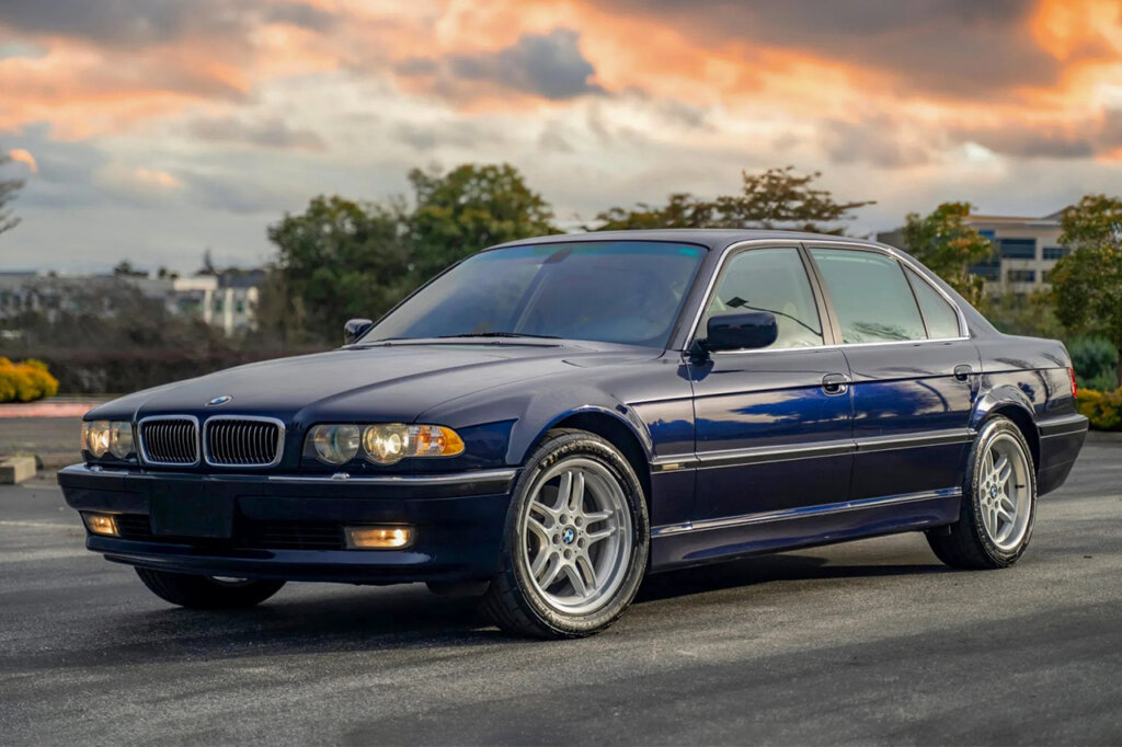 Blue BMW 740iL driving on the road with orange clouds during sunset with trees in the background.