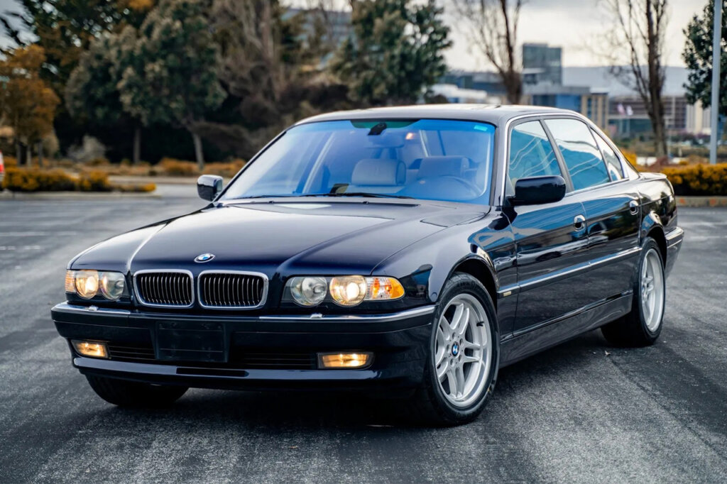 Blue BMW 740iL parked with headlights on with trees and sky in background.
