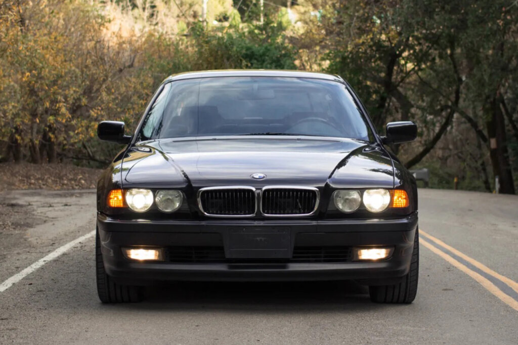 Front end shot of a BMW 740i with headlights and fog lights on parked on a road with trees in the background.