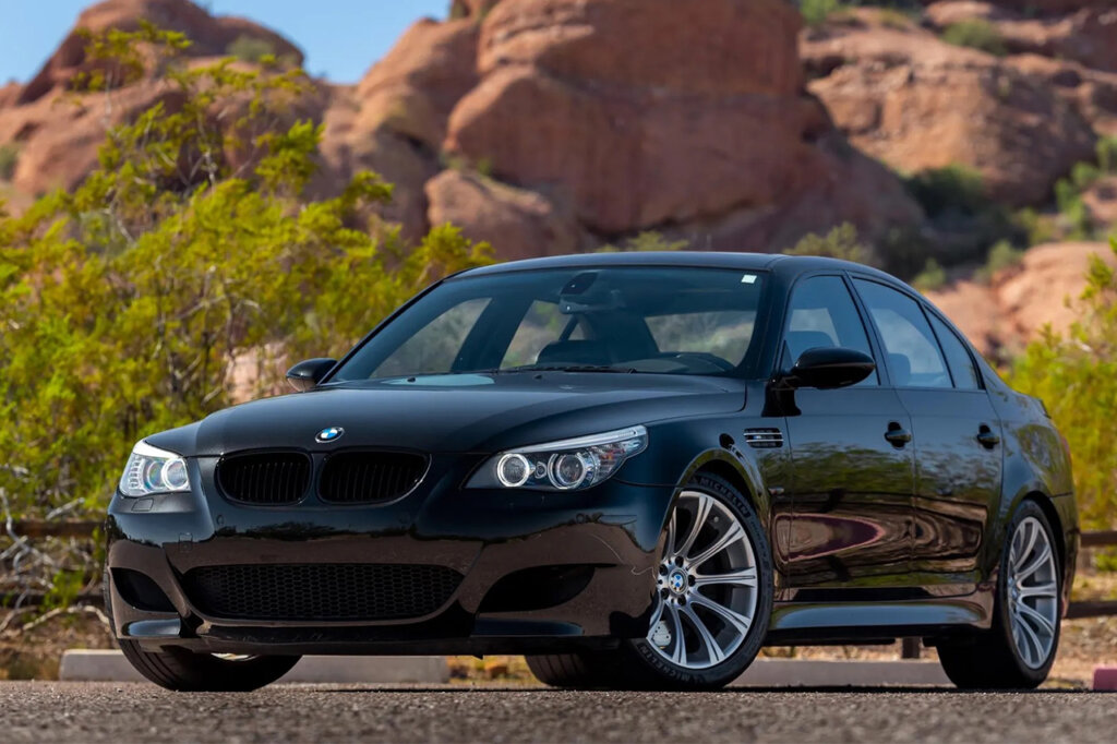 Black BMW M5 E60 saloon parked with wheels turned in front of a rocky cliff and trees in background.