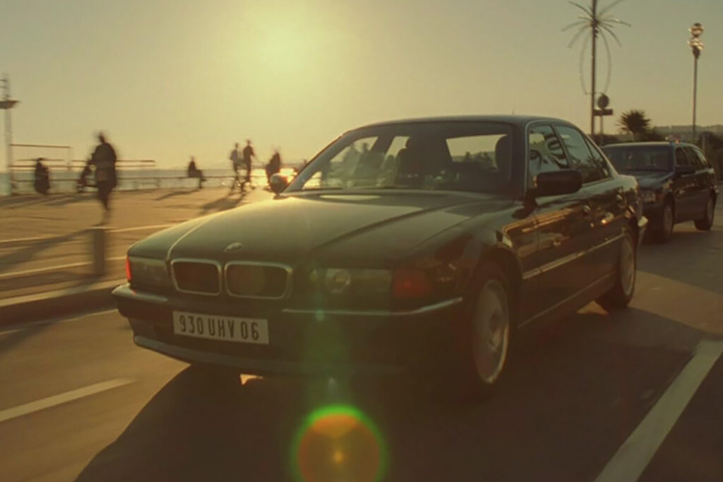 Black BMW 735i sedan driving on a road with traffic, people walking and sun setting in the background.