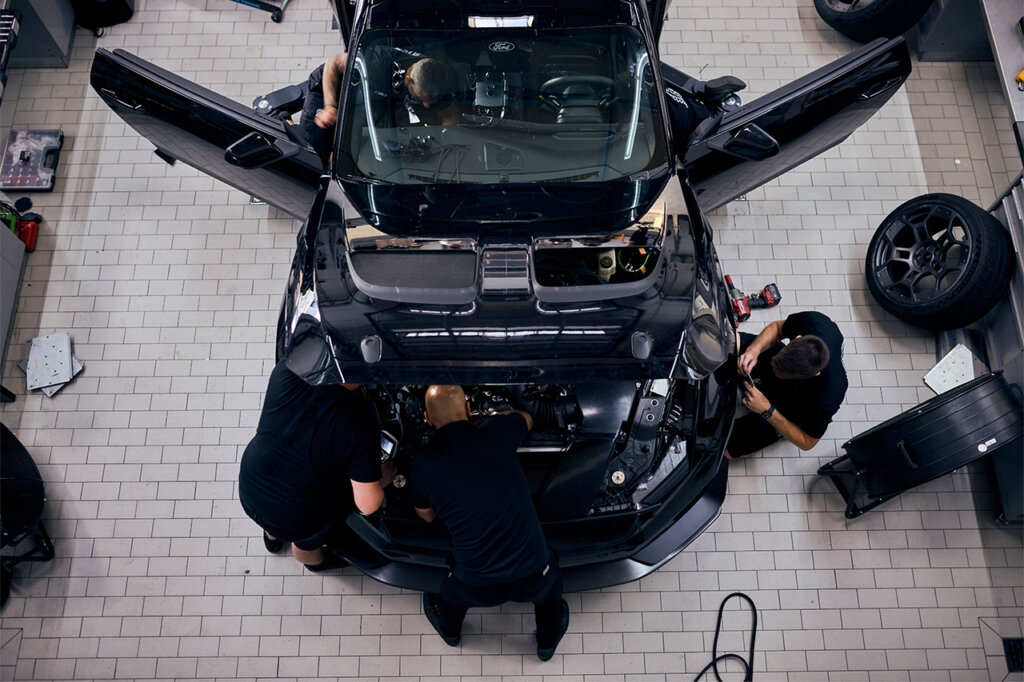 Aerial view of several people in black shirts at the front of the Mustang GTD with the hood removed exposing the engine. Both doors are open with people stretching into it.