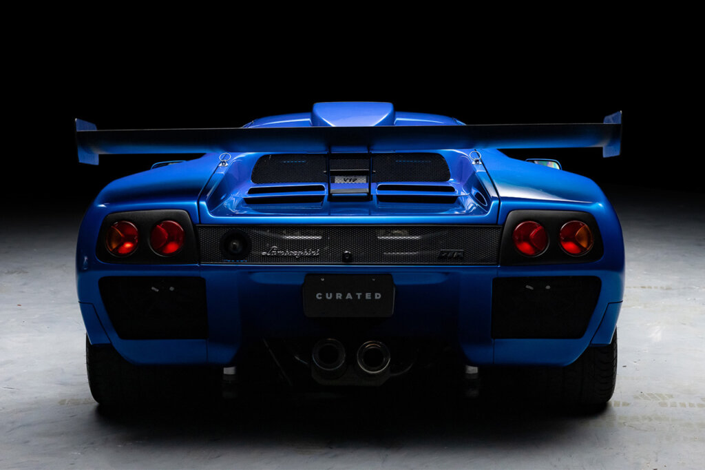 Blu Monterey Diablo GTR rear end with large red taillights with a large fixed rear spoiler above them. Car is shot with a black background and concrete floor.