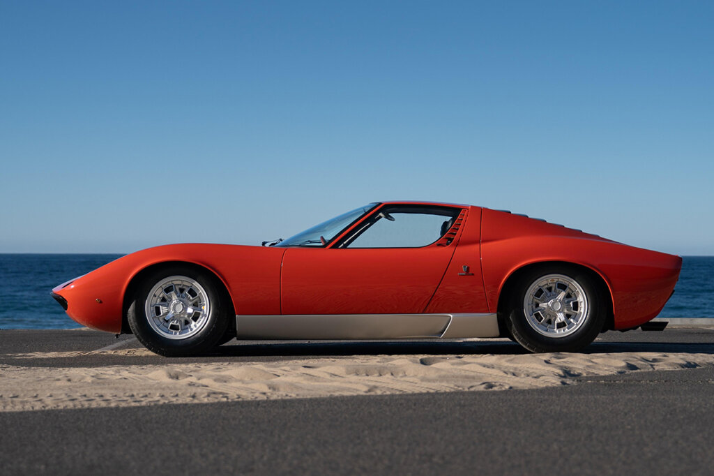 Red Lamborghini Miura P400 side profile parked on sandy road with blue ocean and clear blue sky in background.