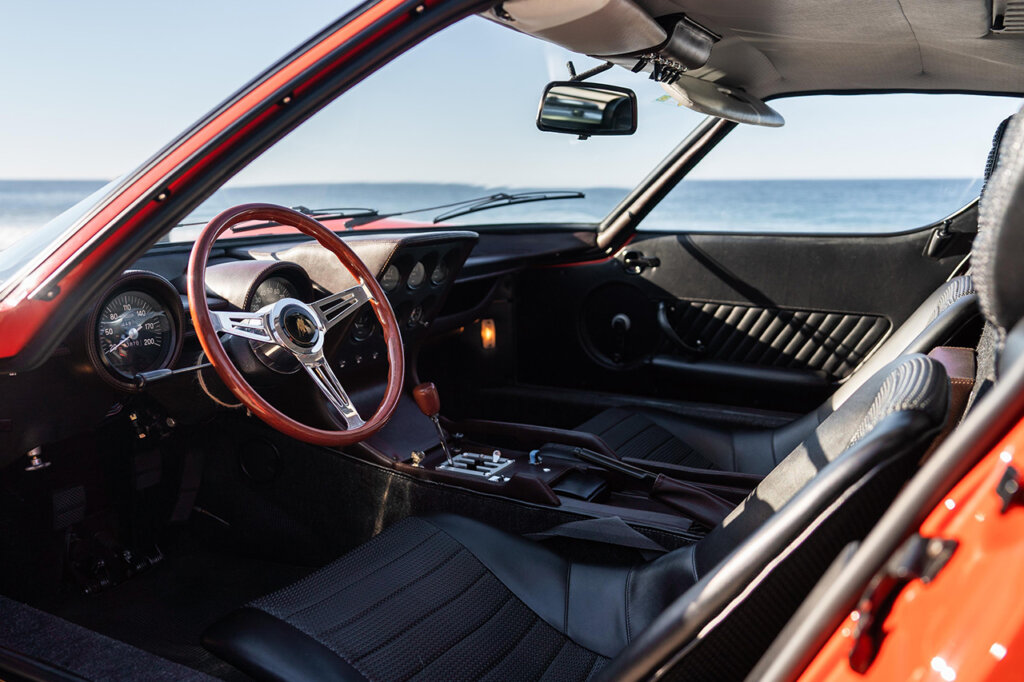 Black leather interior with wood grain steering wheel of a red Lamborghini with the ocean in the background.