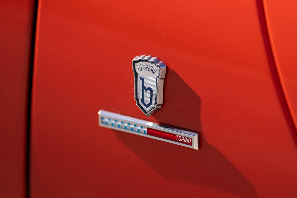 Chrome badging that is the logo of Bertone design above a white and blue checker and red strip on a red Lamborghini car.