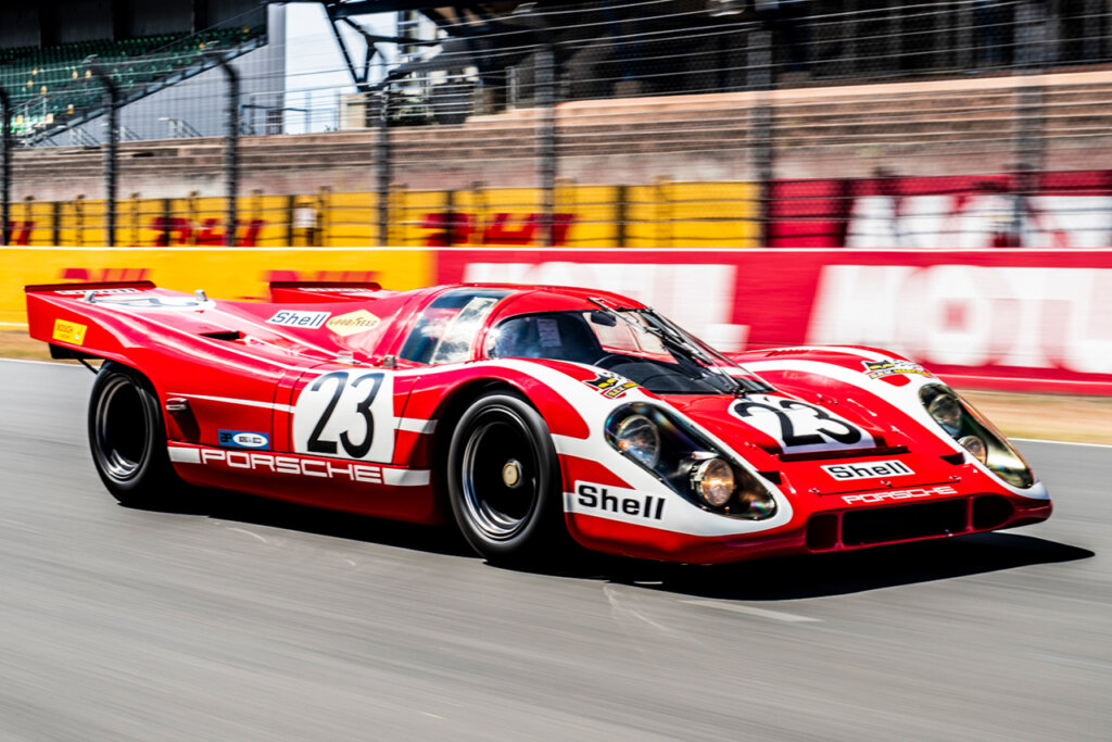 Red #23 Porsche 917K driving fast on a racetrack passing spectator and track barrier.
