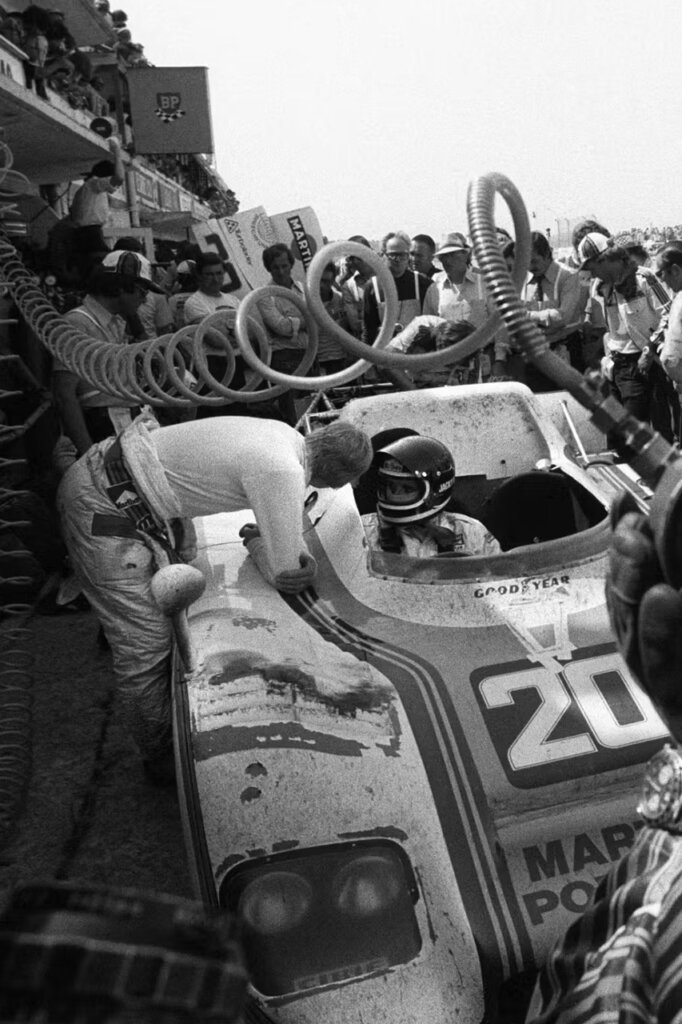 Black and white photo of a race car driver talking to pit crew with many people around the car.
