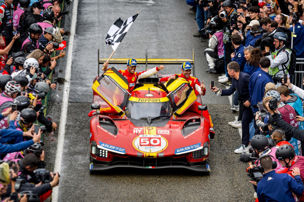 Color photo of a red and yellow hypercar as it crosses the finish line closely surrounded by spectators and the driver is holding a checkered finish flag.