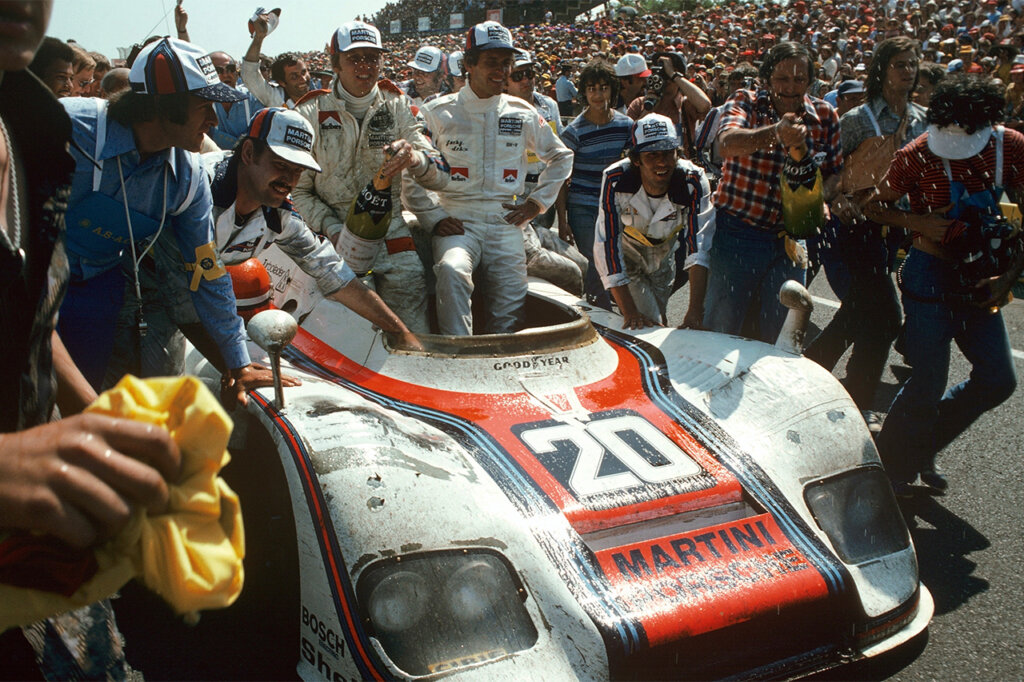 Race car drivers sitting on top of the Porsche 917 Martini race car after winning a Le Mans race closely followed by many spectators and crew.