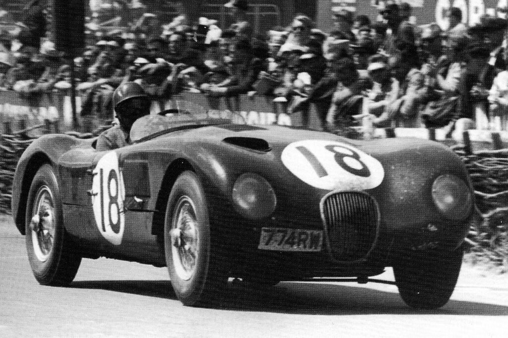 Black and white photo of a Jaguar D-type number 18 passing a crowd of spectators behind the car during 24 hours of Le Mans race.