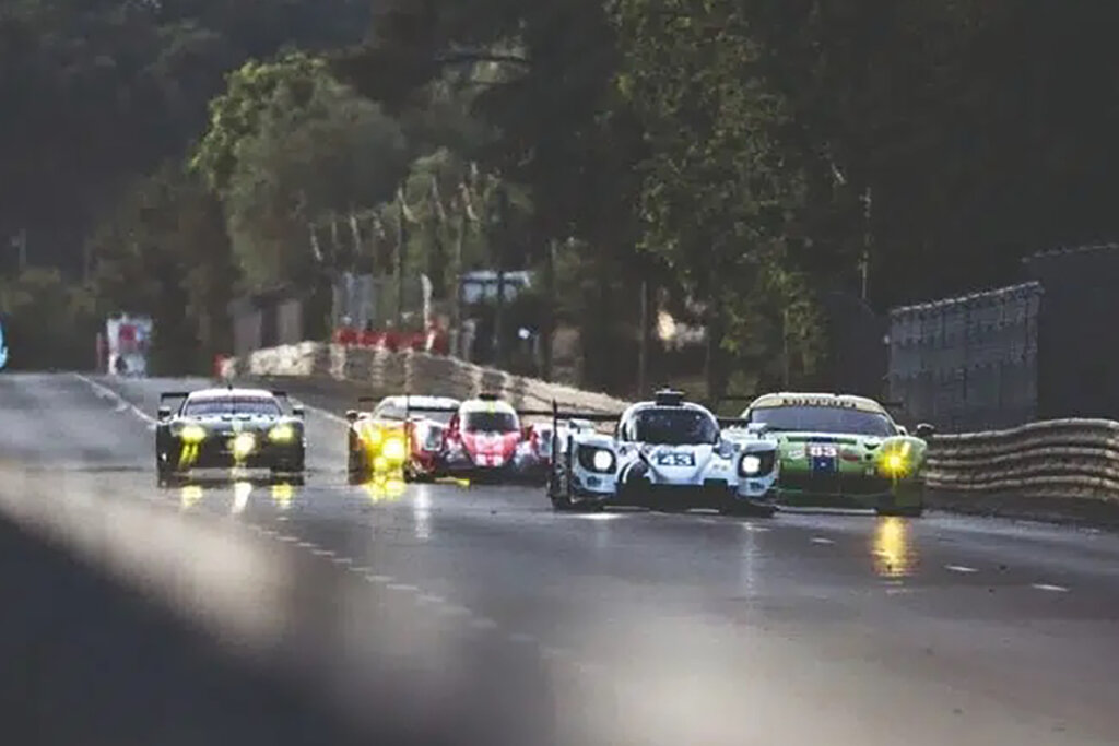 race cars next to one another on the Mulsanne Straight driving fast with lights on passing trees.