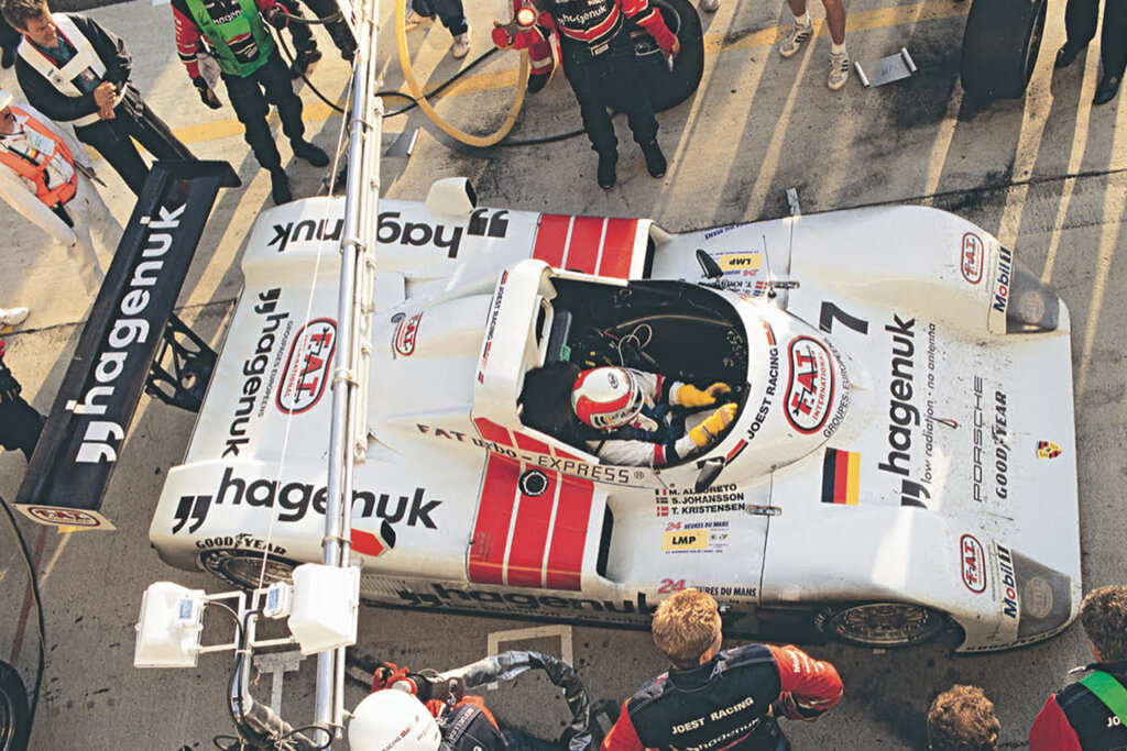 Race car driver in a single cockpit Porsche race car ready to go in the pits surrounded by several people.