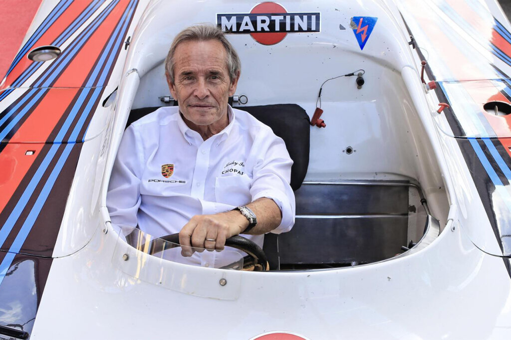 Color photo of a man in a white shirt behind the wheel of a single cockpit Martini Livery Porsche 917 smiling.