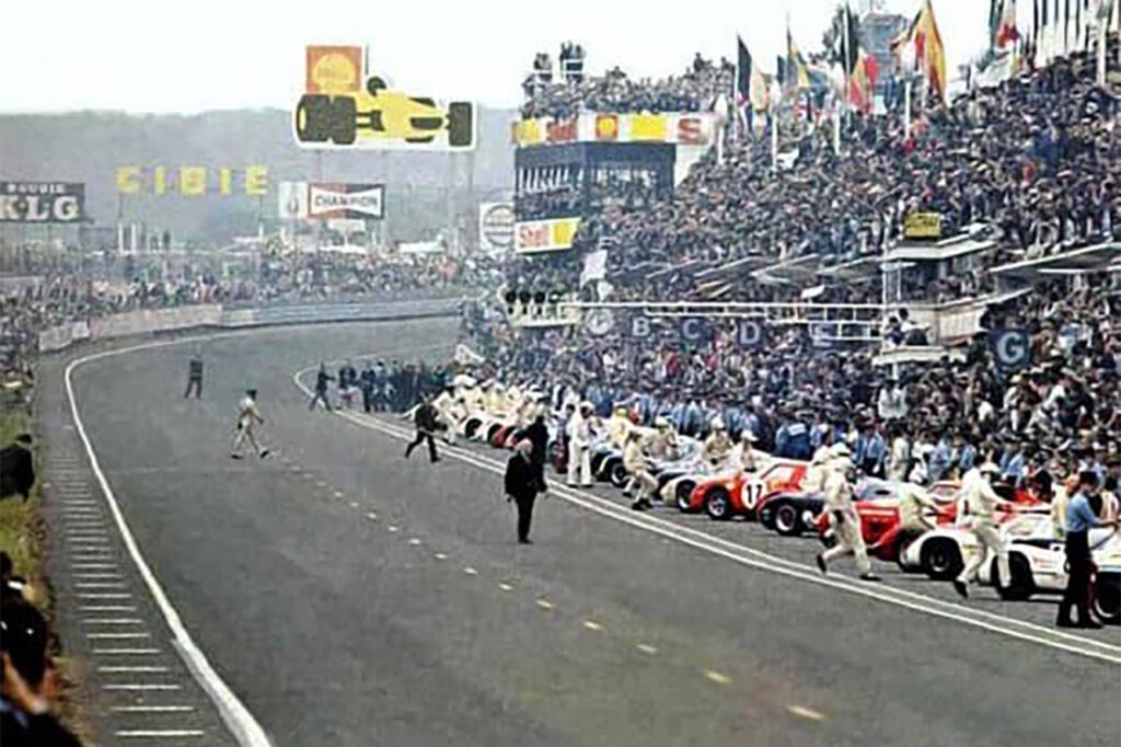 Wide photo of race cars to the right and people on the race track, with crowd of spectator seated in the background.