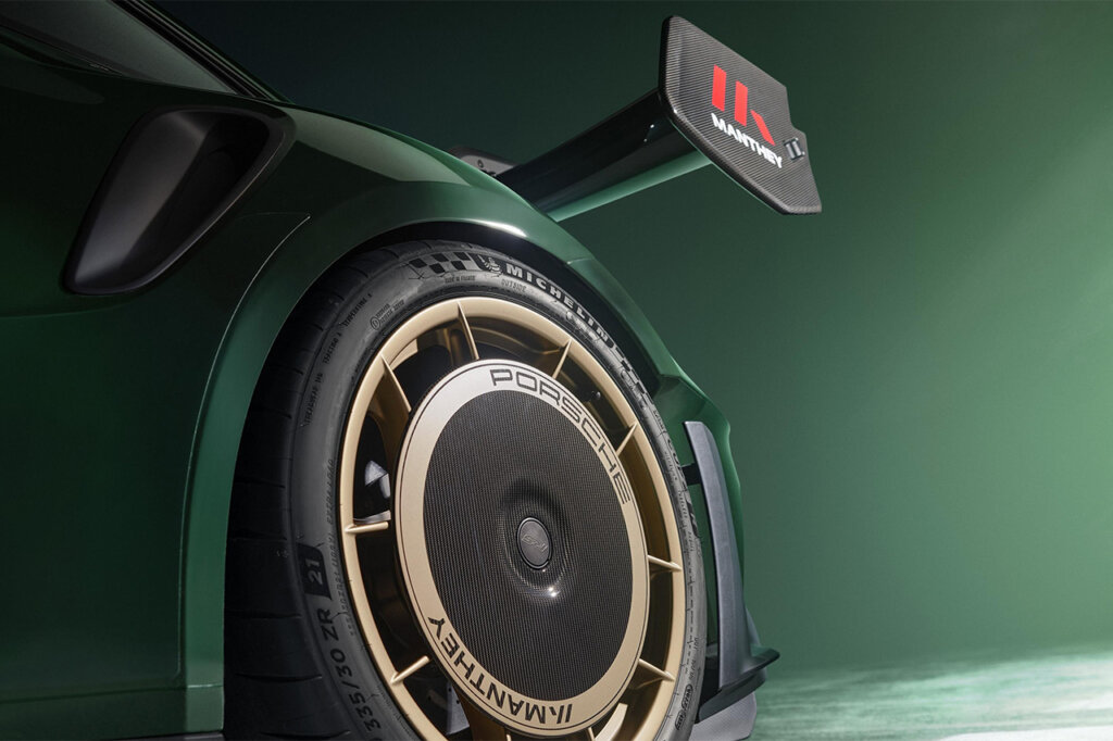 Bronze wheel on a green Porsche 911 with large rear wing with Manthey Racing decal on the end of the spoiler, with a green wall in the background.