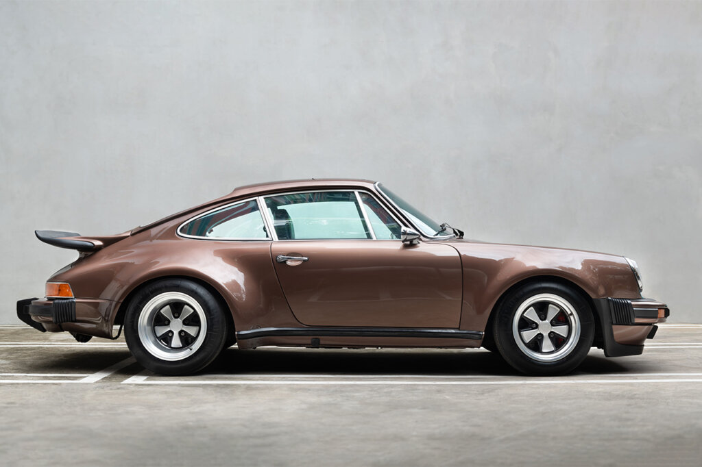 Brown 911 930 Porsche parked with chrome 5 spoke wheels with a solid concrete wall behind it