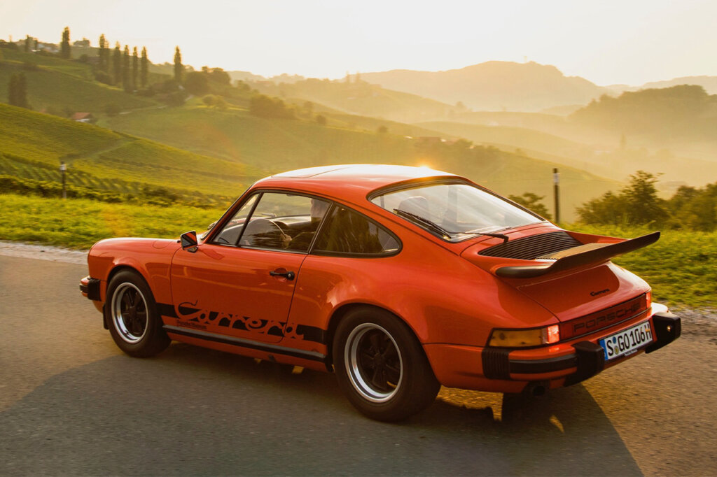 Orange and black 911 Carrera parked on the side of the road overlooking mountains with the sun in the background