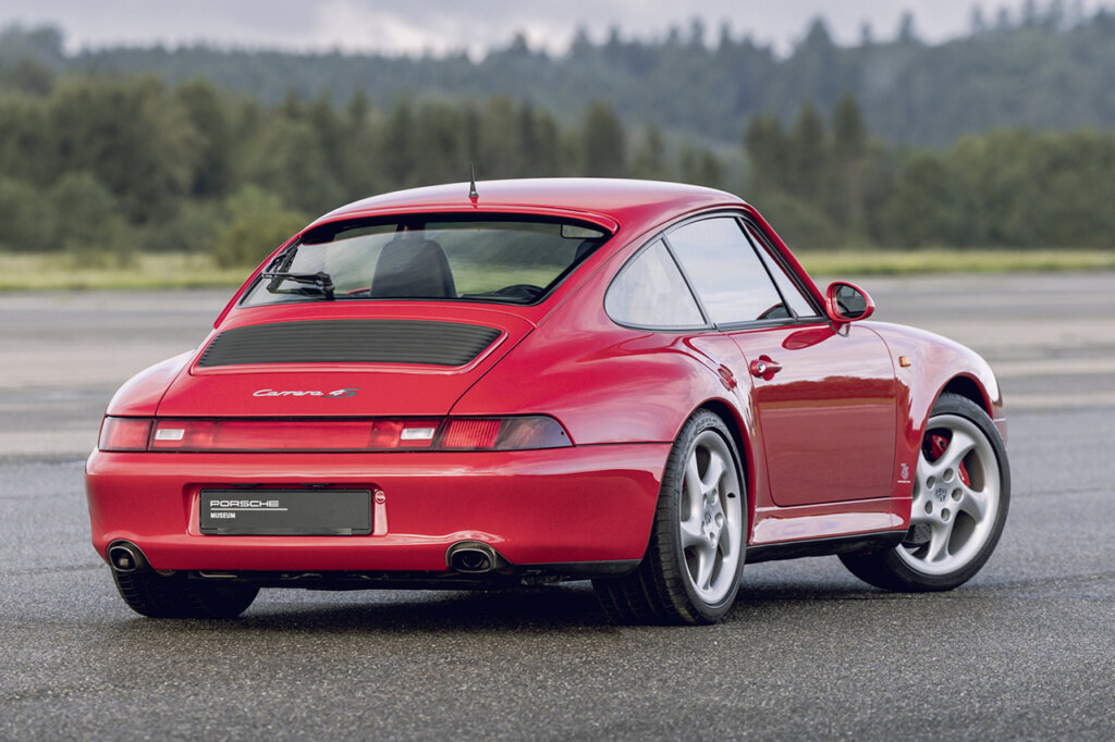 Pink 911 993 parked on asphalt with grey clouds and trees in the background