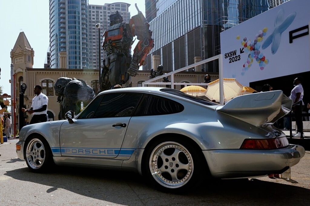 Silver Porsche 930 Whale Tail vehicle with polished chrome wheels and blue accents where the car is parked on a street with city skyscrapers and replica Optimus Prime model in the background.