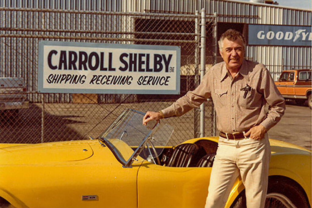 Man standing next to a Yellow Shelby car in front of a fence that has Carroll Shelby business logo behind him on the fence.