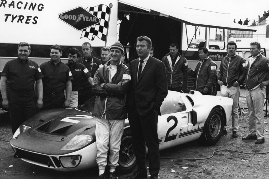 Black and white photo of two men standing next to each other in front of a Ford GT40 with men surrounding the car as well in the background.