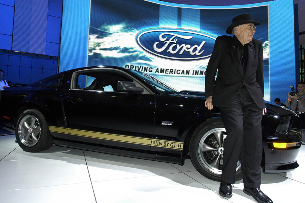 Older man in a black suit with a cowboy hat standing in front of a black and gold Ford Mustang with Ford logo behind it.
