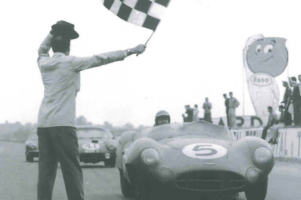 Black and white photo of a man holding a checkered racing flag with a race car and a man driving it with a helmet and number 5 on the hood with other cars and people in background.