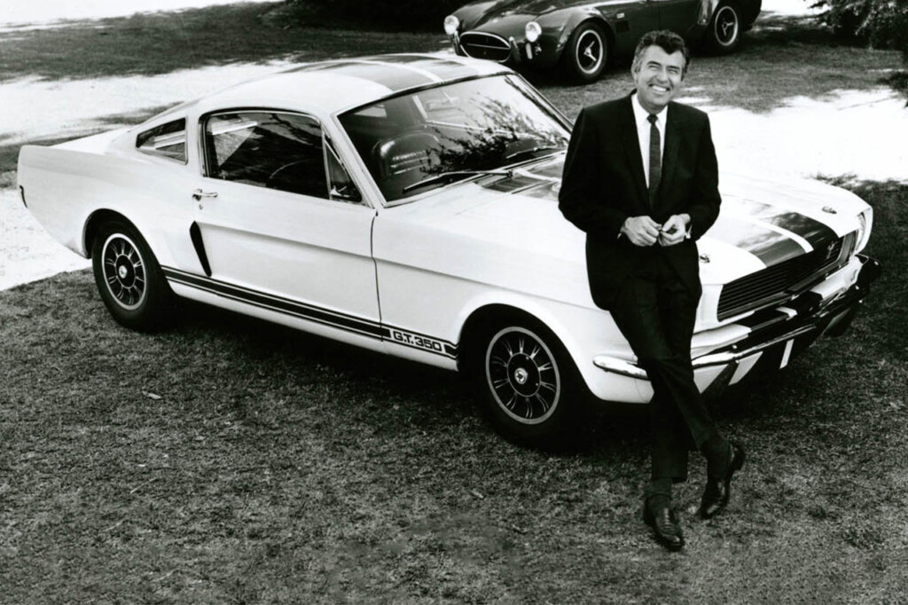 Black and white photo of man smiling in a tuxedo leaning on the hood of a White Ford Mustang GT350 parked on the grass.
