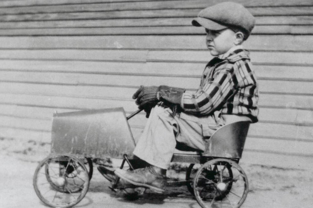 A black and white photo of a kid in a pedal tricycle with a wooden fence in background.