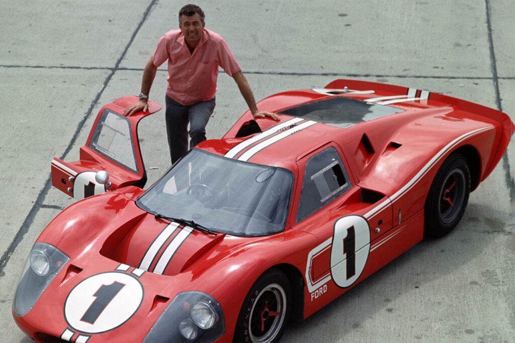 Man in red shirt standing next to an open passenger door of a red Ford GT40 with racing stripes and number 1 on hood and door.