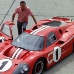 Man in red shirt standing next to an open passenger door of a red Ford GT40 with racing stripes and number 1 on hood and door.