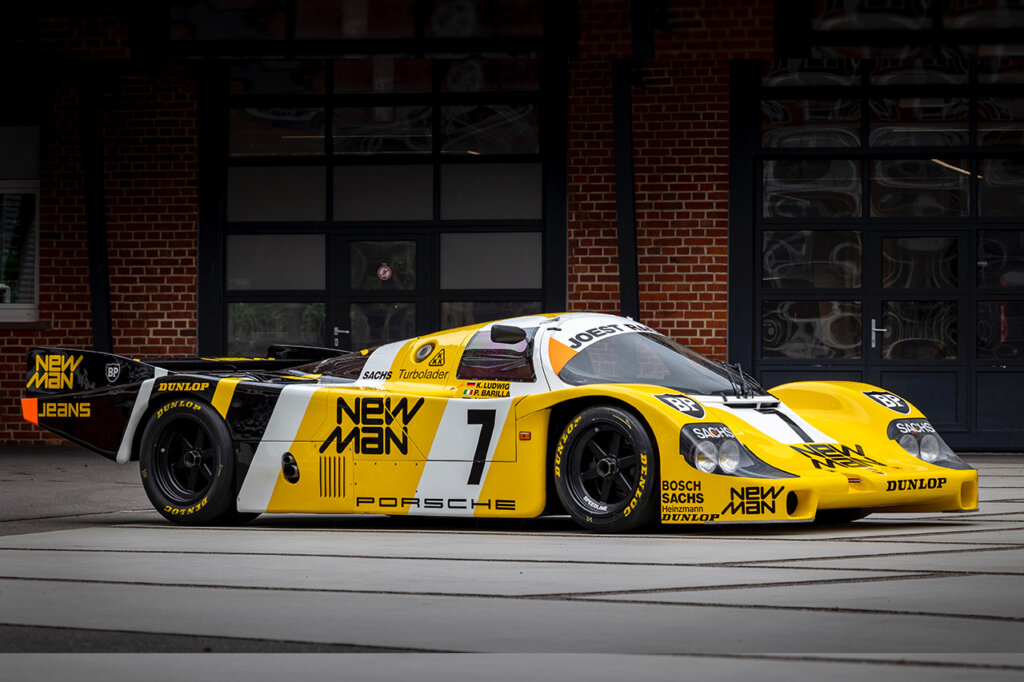 Yellow Porsche Joest Racing 956LH race car parked in front of a building with windows behind it and concrete in the foreground.