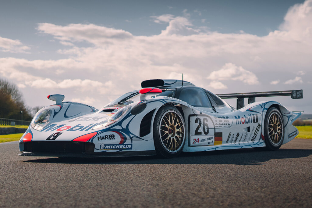 White with red and blue Porsche 911 GT1 with large fixed rear spoiler and roof scoop air induct parked at an angle on a road with cloudy blue sky in background.