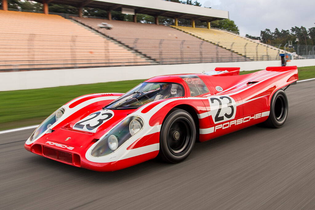 Red Porsche 917KH race car with man in single seat cockpit driving on the racetrack passing empty audience stands.