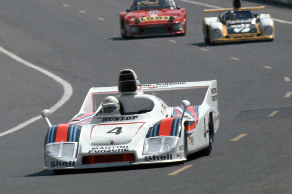 White Porsche 936 Martini race car #4 with a single driver with a white helmet in front of other race cars on a track.