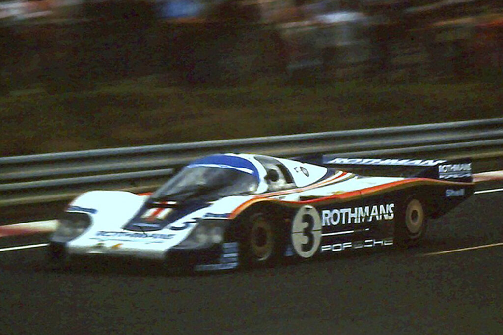 White, Blue Rothman Porsche 956 LH driving through a turn on a race track with a metal barrier and trees in the background.
