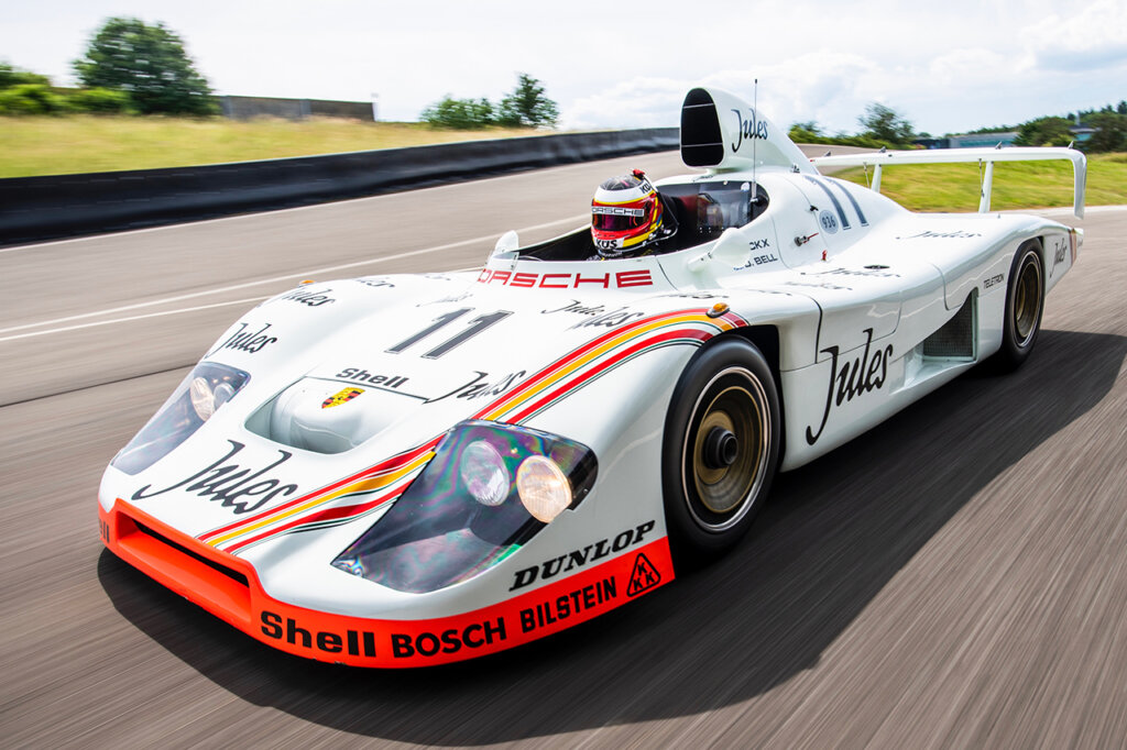 White Porsche 936 Spyder with White Jules race livery, orange accents and striping down the front end with driver in the single open cockpit with large air duct. The car and driver are driving down a track with grass and tree and blue sky in background.