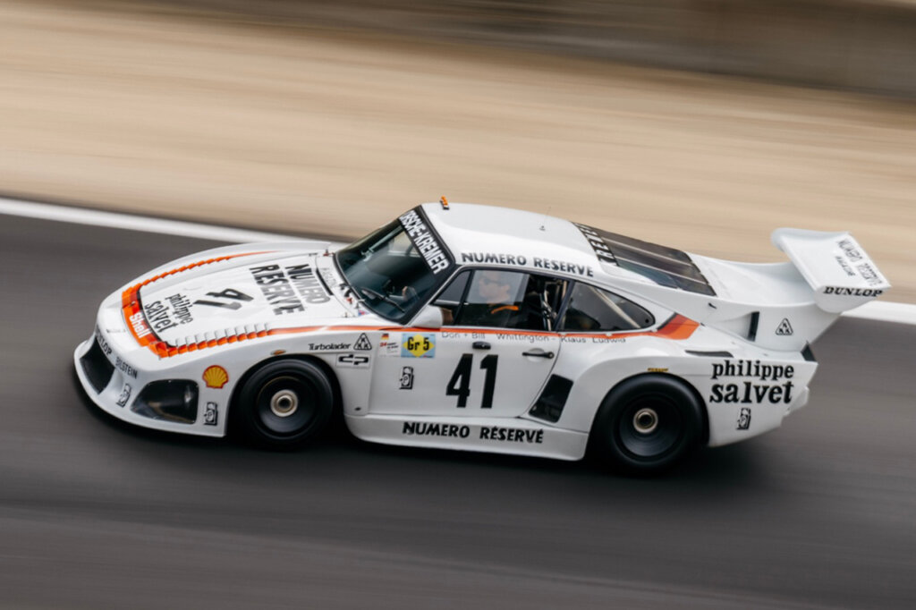 White Porsche 935 with orange striping from the hood to the door panels with various stickers, branding and number on door panel with the car and driver on a race track.