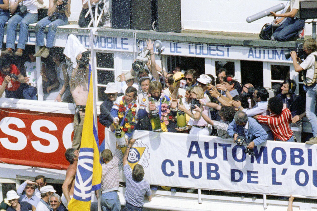 Crowd of people surrounding race car drivers being sprayed with champagne, people cheering with banners win red and white with letters on it.