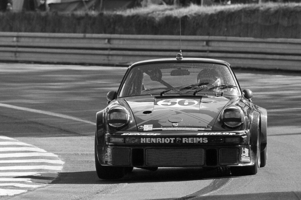 Black and white photo of a Porsche 911 with a driver in a white helmet driving through a turn on a racetrack.