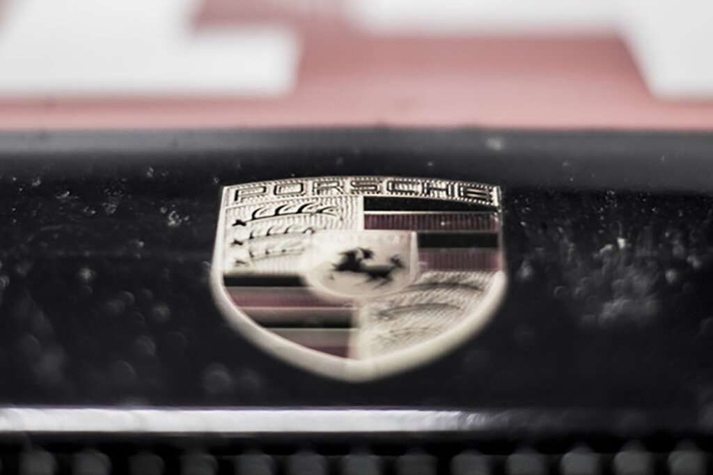 Porsche Crest in the foreground with blurry out of focus closeup of a car grill and a red background with white lettering.