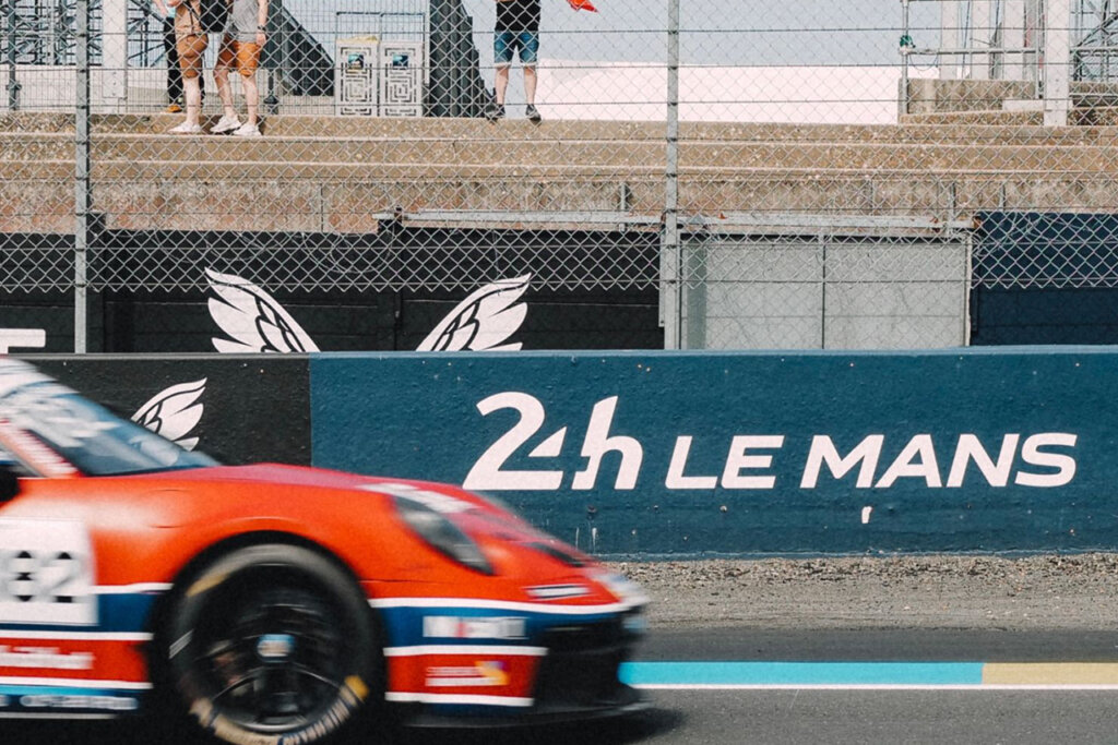 Red Porsche 911 with decals and branding in the foreground passing a blue divider with 24h Le Mans branding in white painted on it.