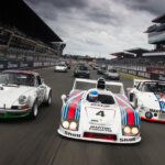 Three white Porsche vehicles in Martini race livery and leading a pack of other cars in the background on a racetrack with grey cloudy sky in background with stadium stands.