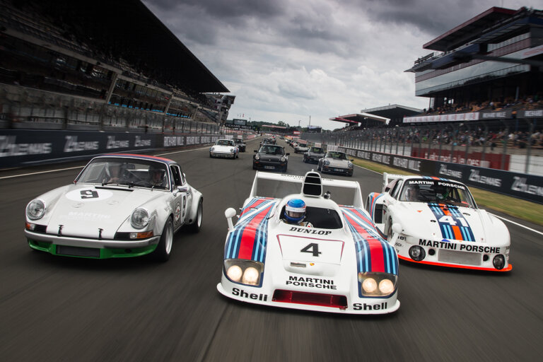 Three white Porsche vehicles in Martini race livery and leading a pack of other cars in the background on a racetrack with grey cloudy sky in background with stadium stands.