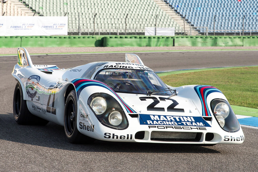 White Porsche 917KH Number 22 Martini race car parked on the race track with empty bleachers in the background and green grass next to it.