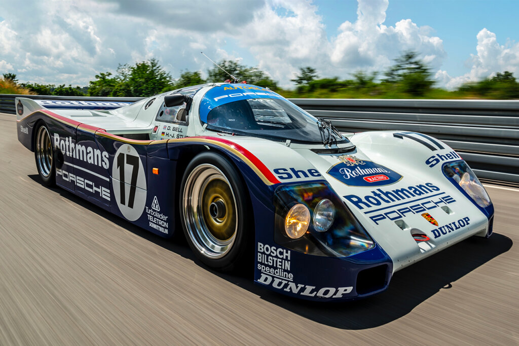 Porsche 962C with Rothman livery driving on road with blue sky and trees in the background with the headlights on.