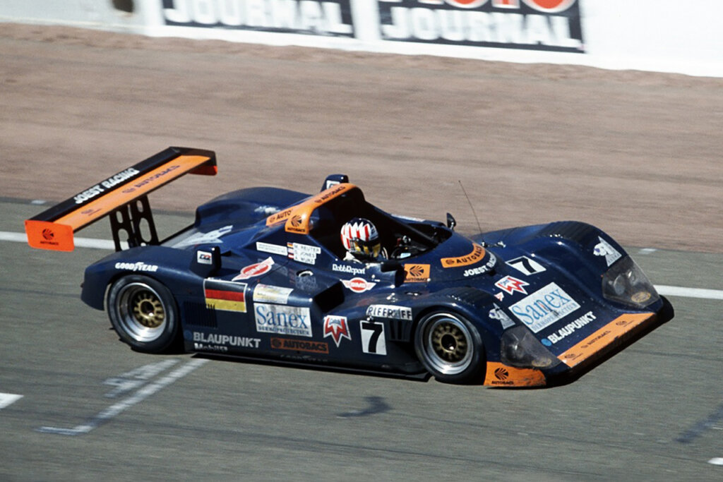 Black open top TWR-Porsche WSC race car with a single driver in the cockpit wearing a helmet on the racetrack passing a white divider with green banners on it.