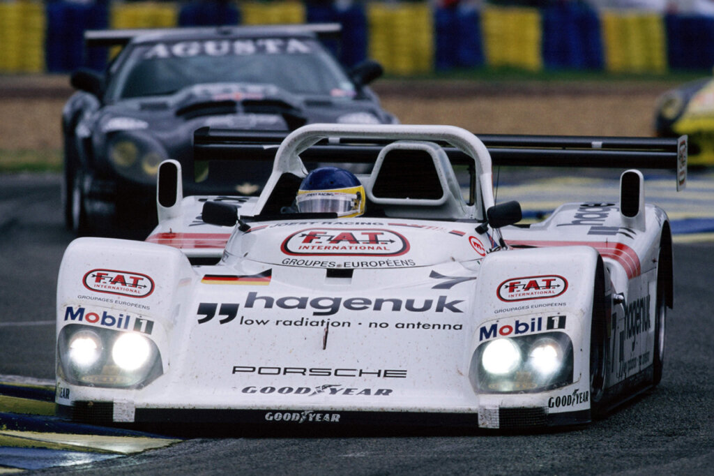 White TWR-Porsche spyder with headlights on driving through a turn with a black race car following closely behind.