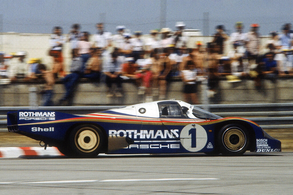Porsche 956LH Rothman livery driving through a turn passing spectators in the background.